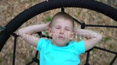 一个蓝眼睛<strong>的</strong>5岁男孩穿着一件蓝色T恤，闭着眼睛做梦，躺在一个<strong>圆圆的</strong>秋千上。 孩子微笑着
